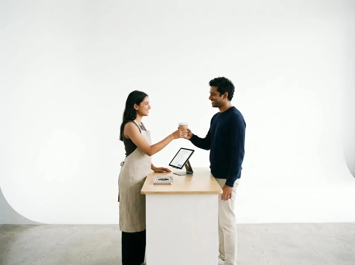 A barista handing a coffee to a regular customer beside a tablet till